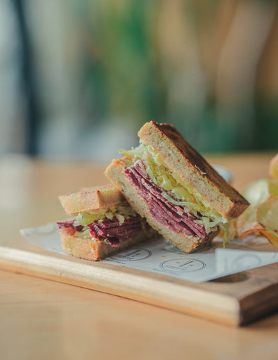 A Reuben Sandwich from Loafology Bakery & Café, filled with corned beef, shredded lettuce, and sauce, is cut in half and served on a wooden board lined with white paper.