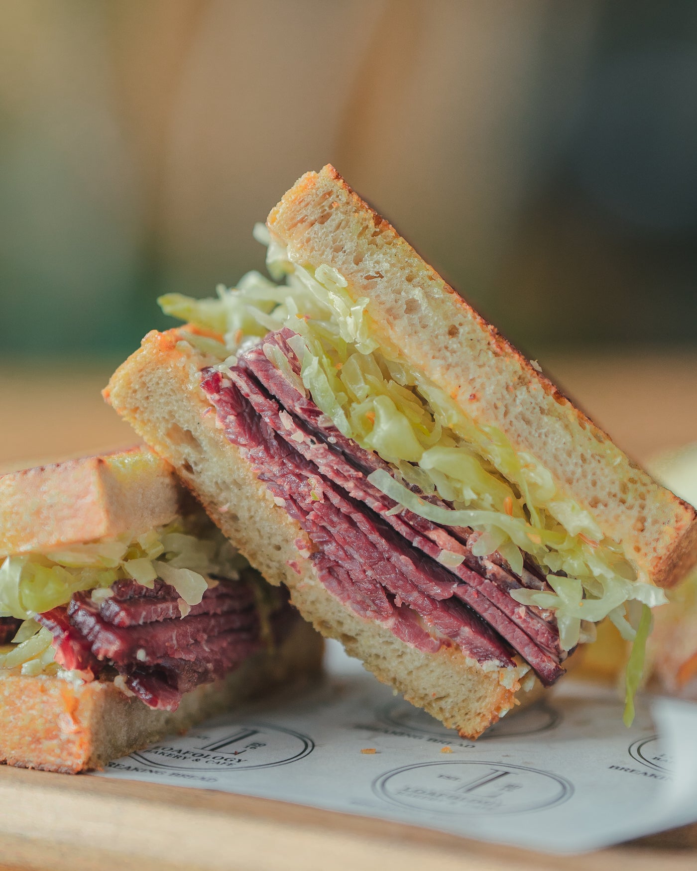 A Reuben Sandwich from Loafology Bakery & Café, featuring layers of meat, lettuce, and condiments on toasted bread, all placed on parchment paper adorned with a circular logo.