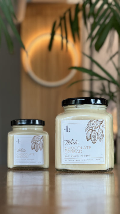 Two jars of Loafology Bakery & Café's White Chocolate Spread, adorned with black lids, are displayed on a wooden surface. The labels, which feature text and an illustration of cacao pods, prominently declare "No Artificial Additives." A blurred, circular light fixture is visible in the background.