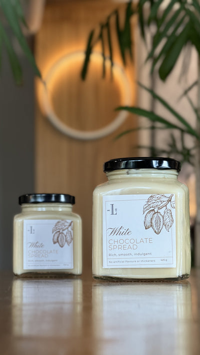 Two jars of Loafology Bakery & Café's White Chocolate Spread placed on a wooden surface, with a blurred circular light and green leaves in the background, reassuringly labeled with no artificial additives.