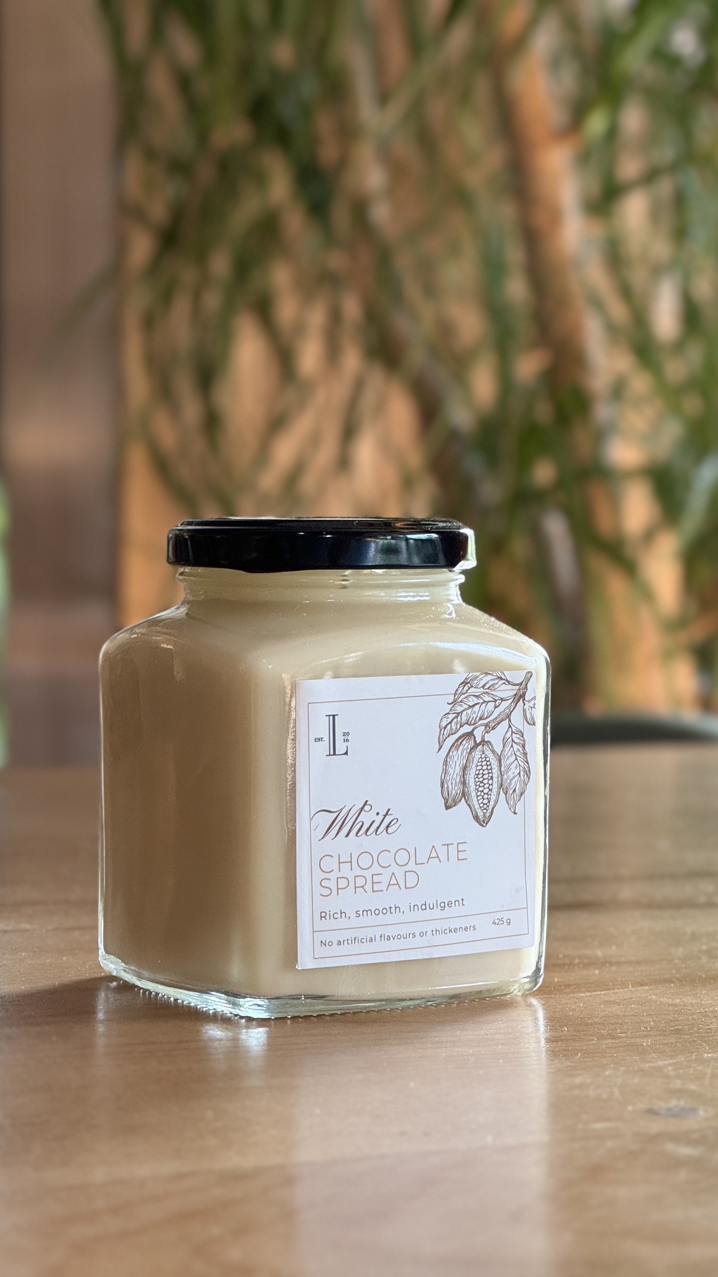 A jar of Loafology Bakery & Café's White Chocolate Spread with a black lid is placed on a wooden table. The label indicates it's rich, smooth, and indulgent, with no artificial flavors or additives.