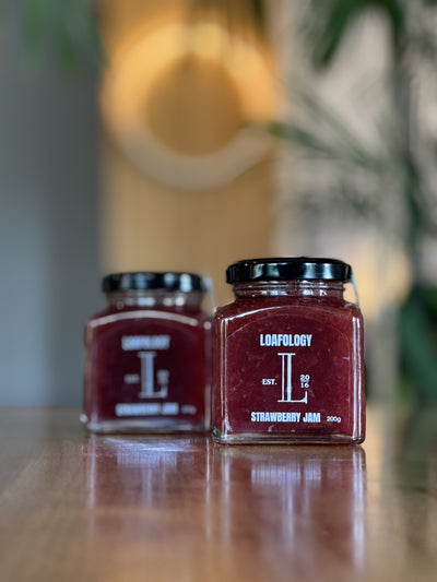 Two jars of Loafology Bakery & Café Strawberry Jam 200g with black lids rest on a wooden surface, showcasing their labels that emphasize no artificial additives, against a blurred background.