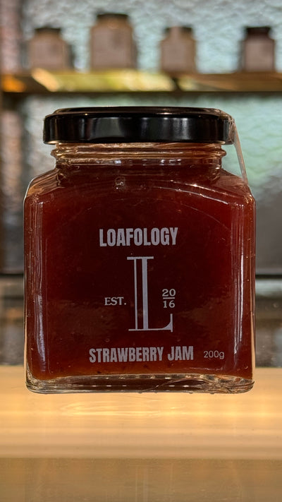 A 200g jar of Loafology Bakery & Café Strawberry Jam, featuring a black lid and a label displaying "Loafology," "Est. 2016," and "Strawberry Jam" with a proud declaration of "no artificial additives," rests on a glass shelf.