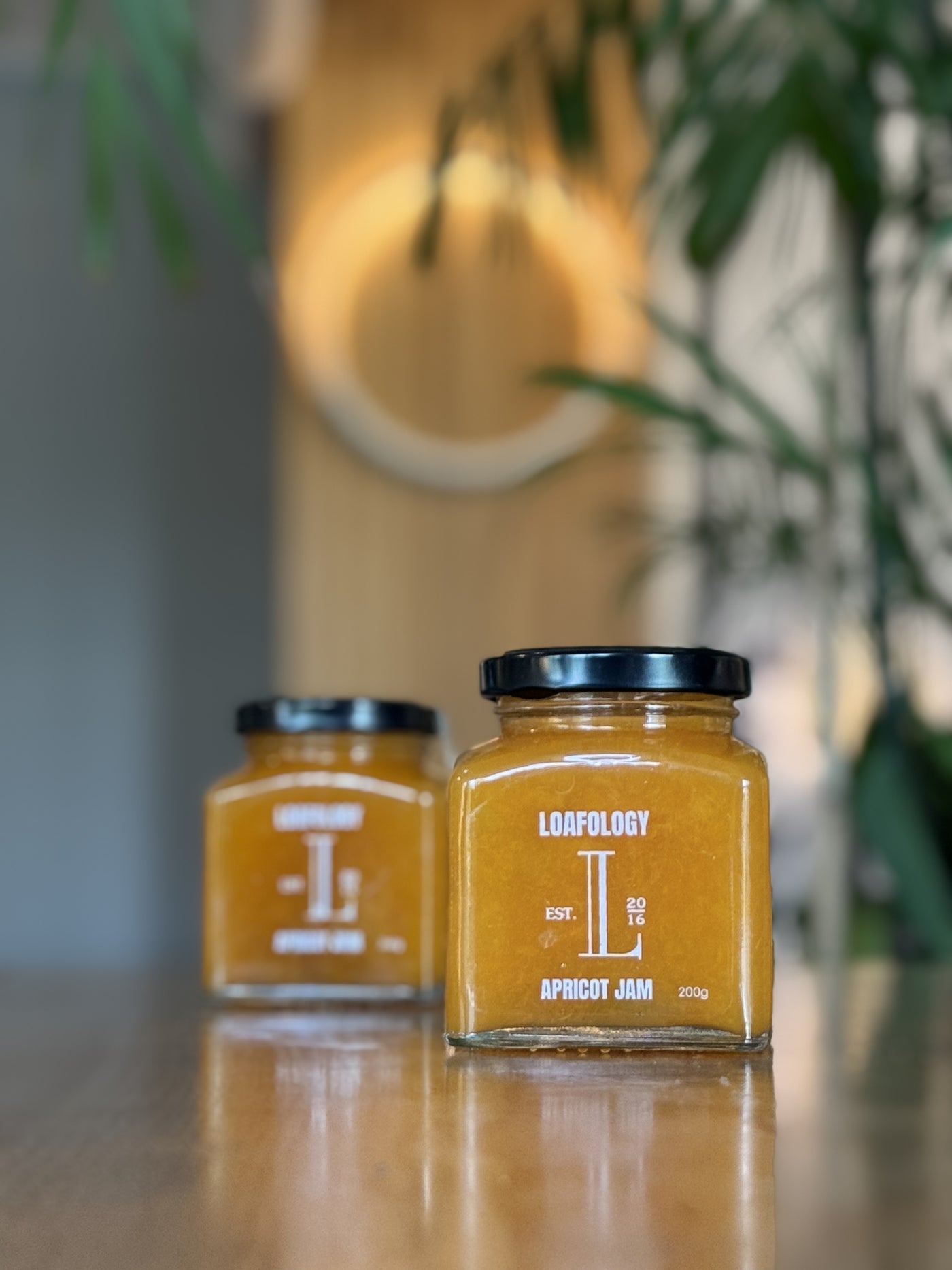 Two jars of Loafology Bakery & Café's Apricot Jam (200g), free from artificial additives, are placed on a wooden surface with a green plant in the background. One jar is in the foreground, while the other is slightly blurred in the background.