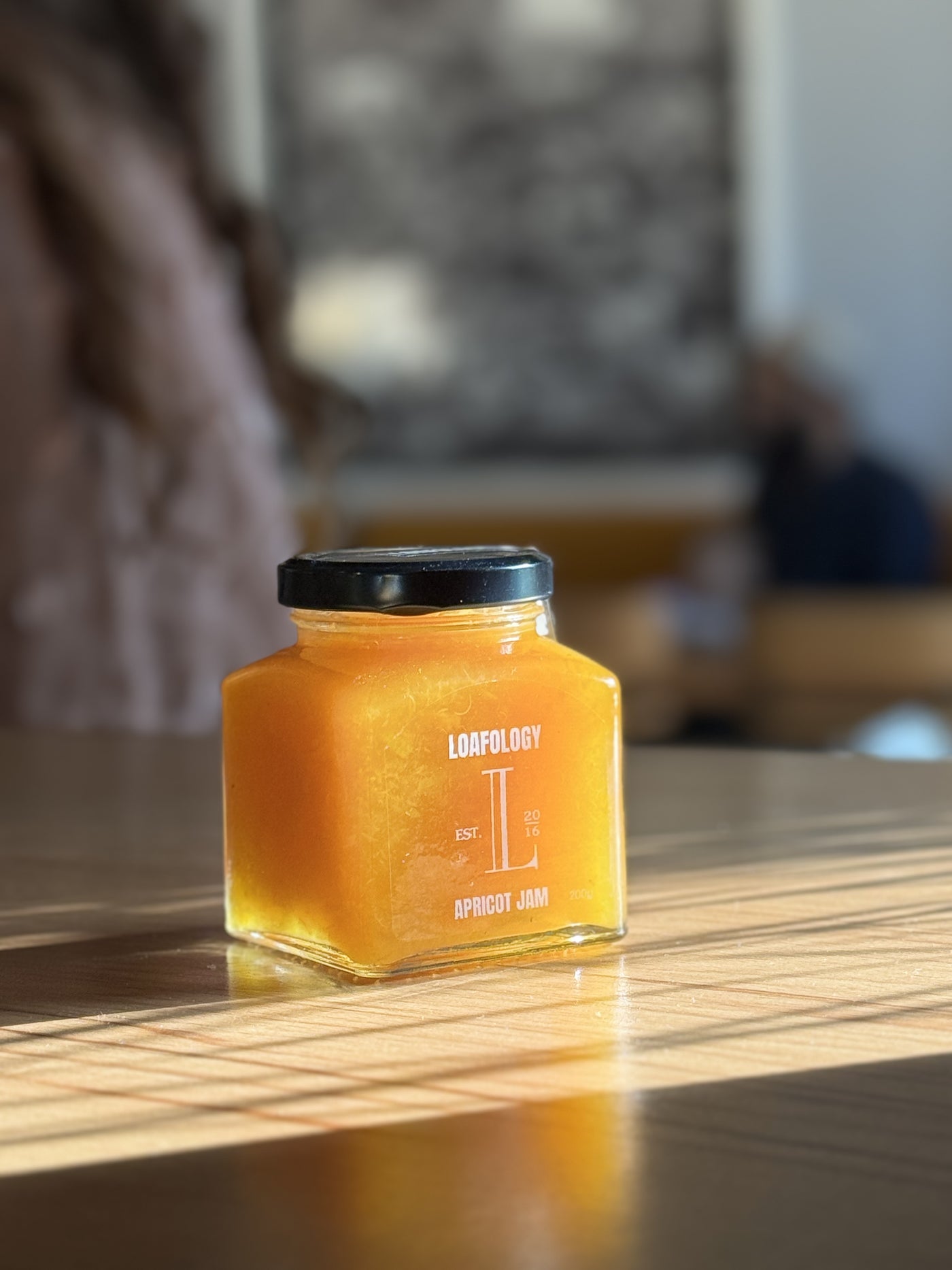 A square glass jar of Apricot Jam 200g from Loafology Bakery & Café sits on a wooden table, with sunlight casting shadows. Proudly made with no artificial additives, it promises pure, natural flavor.