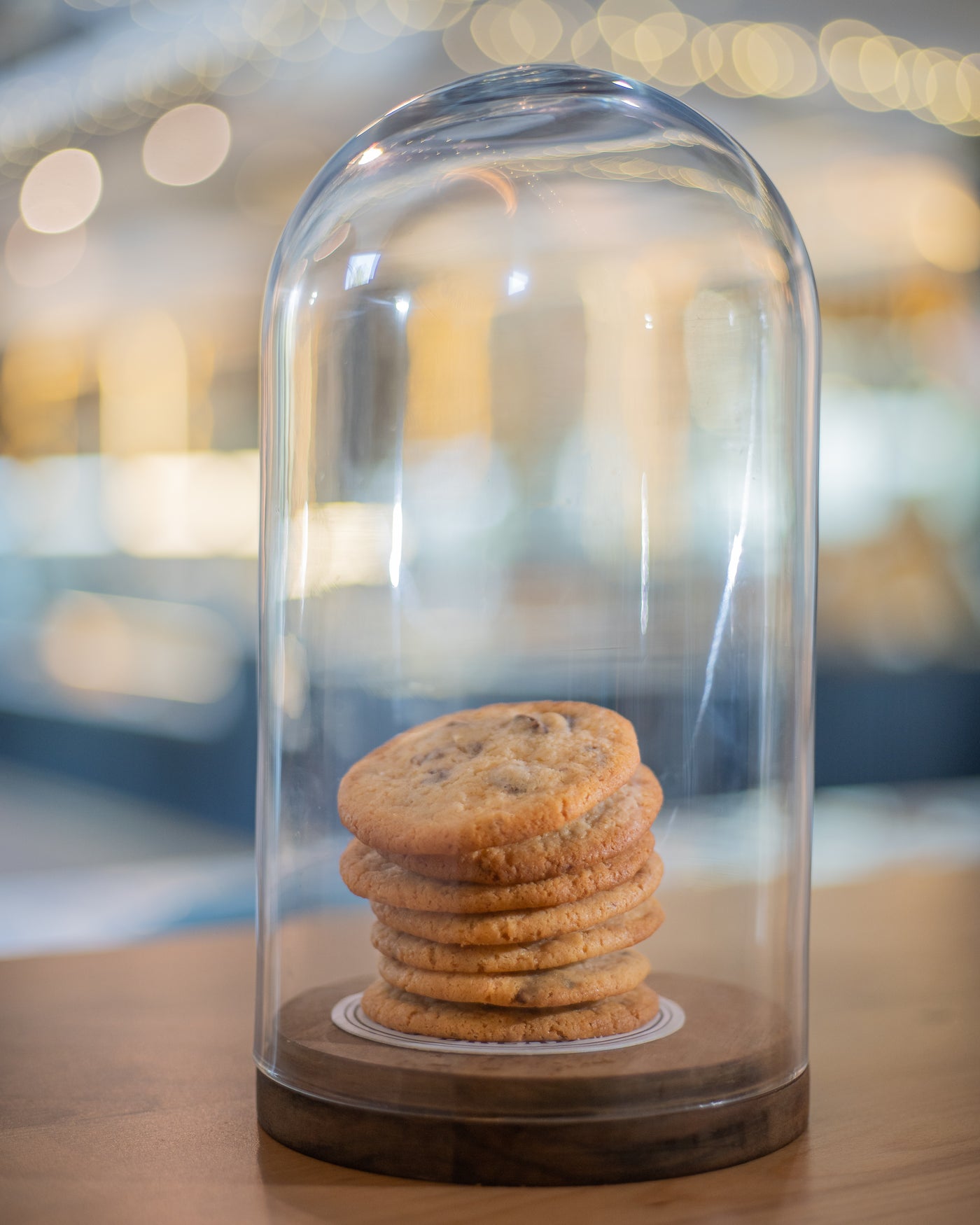 A stack of seven Loafology Bakery & Café Chocolate Chip Cookies is displayed under a glass dome cover, sitting on a wooden base, with a blurred background.