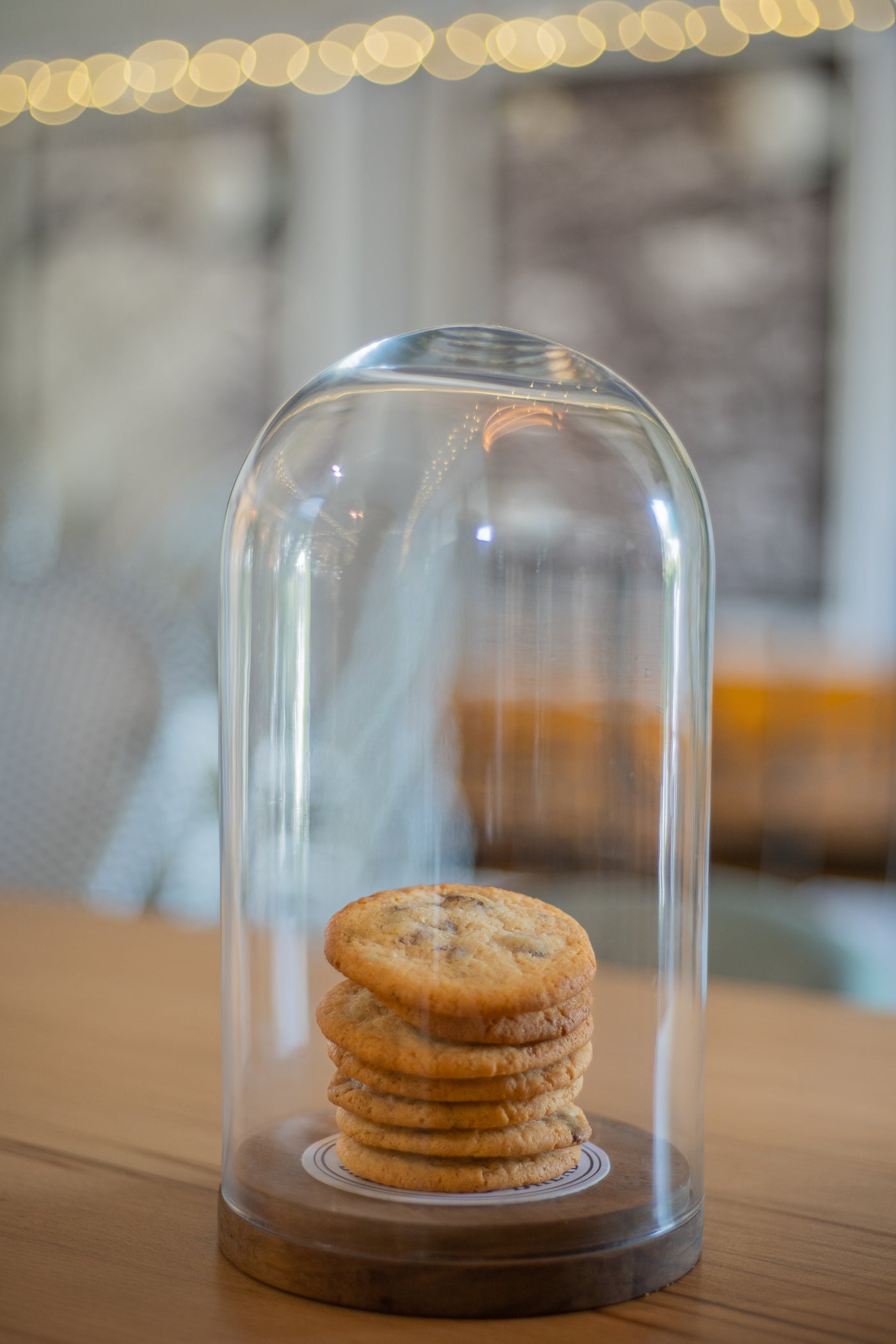 Displayed under a glass dome on a wooden table, a stack of Loafology Bakery & Café's Chocolate Chip Cookies - 7 Pieces stands out against the blurred background.