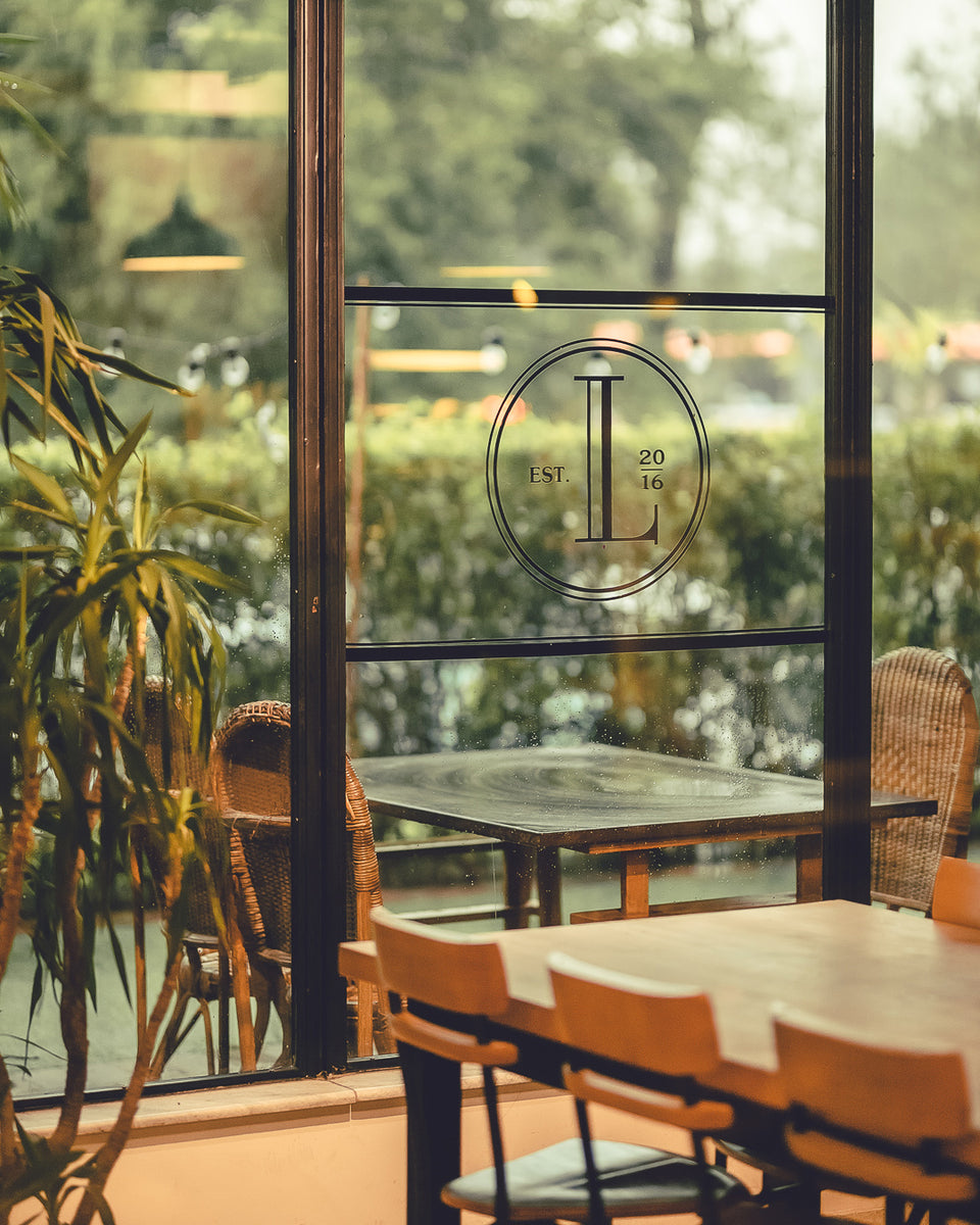 Interior view of Loafology Bakery & Café with wooden chairs and tables near a glass window displaying a logo with "L" and "Est. 2016." Outside, greenery can be seen through the window.