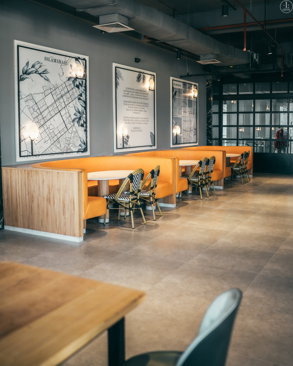 A modern restaurant interior at Loafology Bakery and Café featuring wooden booths with orange seating, black-and-white framed maps on the wall, and stylish chairs with black-striped upholstery.