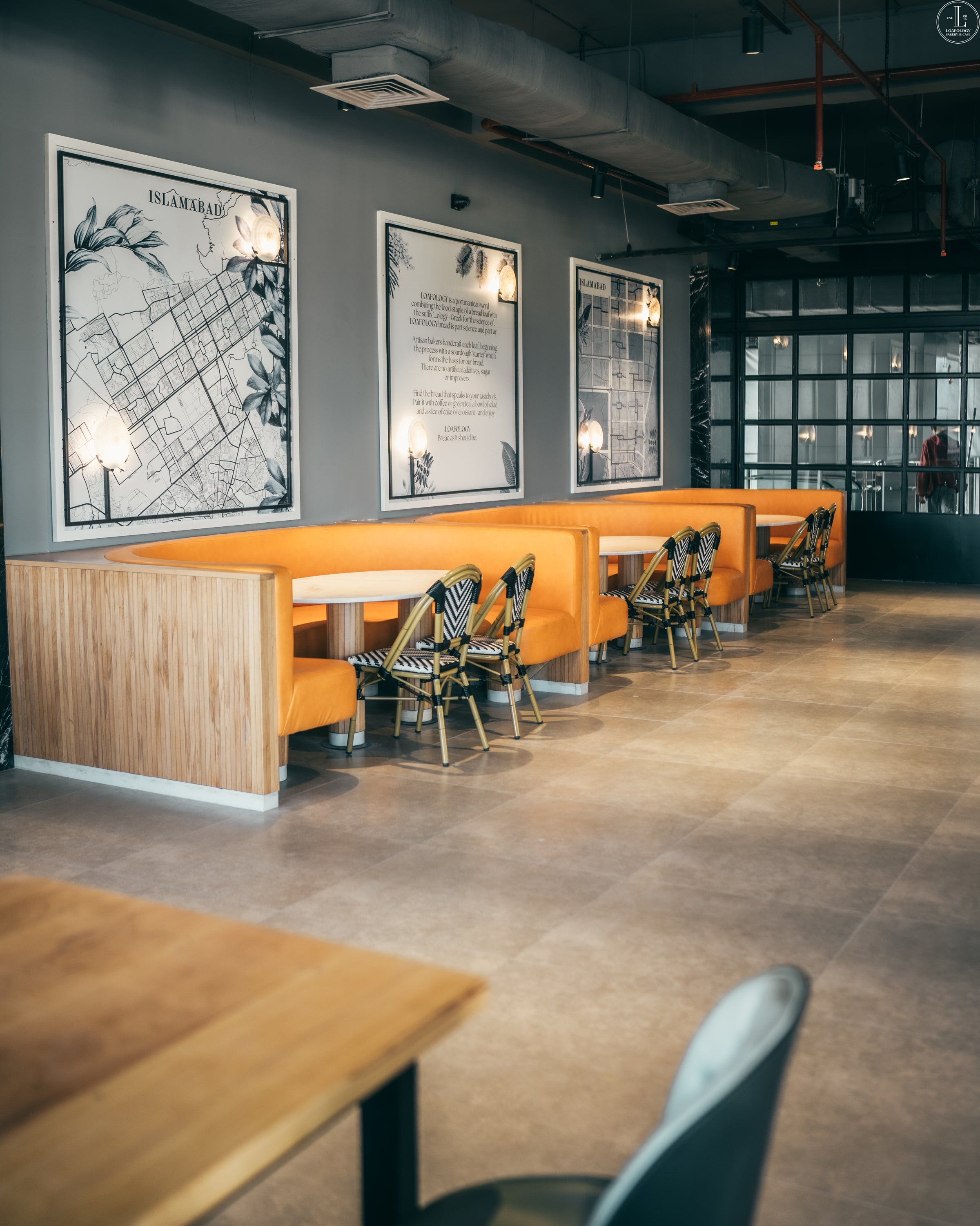 A modern restaurant interior at Loafology Bakery and Café featuring wooden booths with orange seating, black-and-white framed maps on the wall, and stylish chairs with black-striped upholstery.