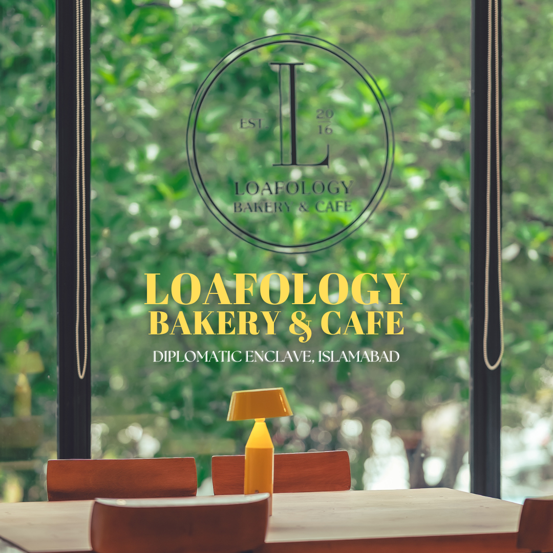 A view of the interior at Loafology Bakery & Cafe in Diplomatic Enclave, Islamabad, showing a wooden table and chairs, with a soft yellow lamp on the table and the cafe's logo on a large window.