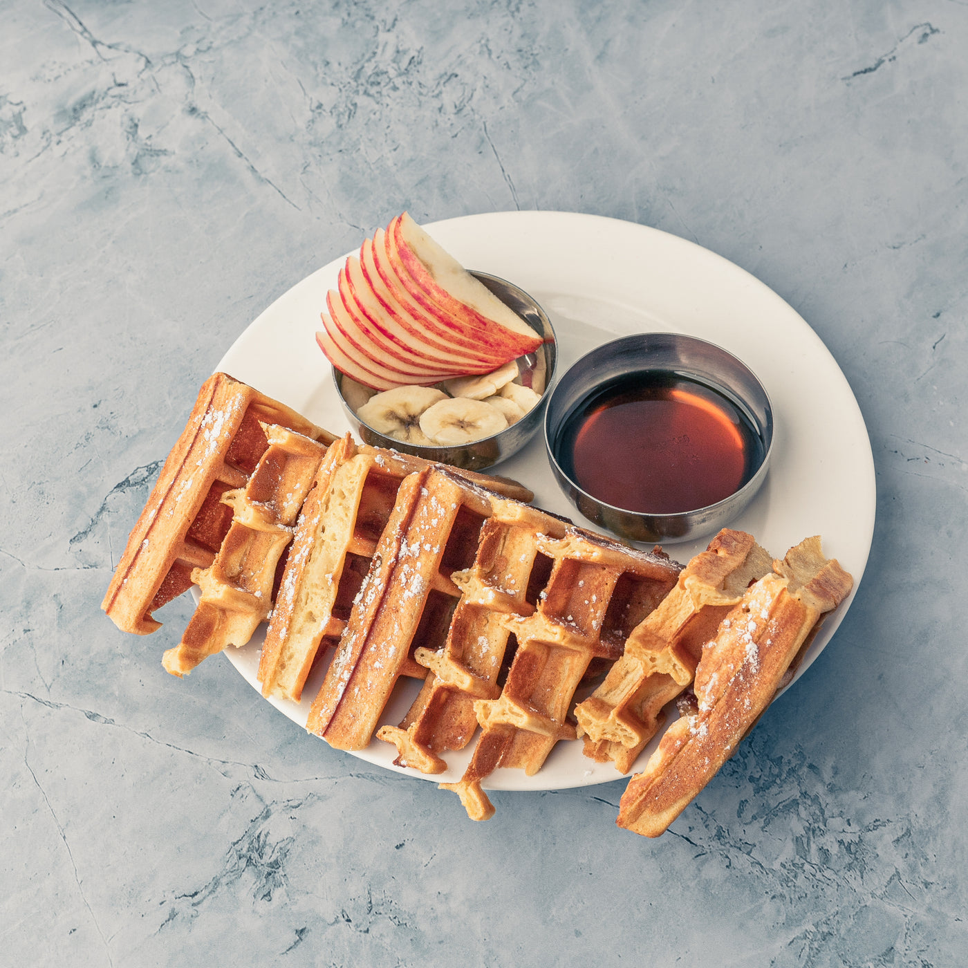 A breakfast delight: Loafology Bakery & Café Waffles dusted with powdered sugar, topped with sliced apples and bananas, accompanied by a small cup of maple syrup on a gray textured surface.