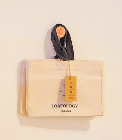 A beige Loafology Canvas Tote Bag (Premium) from Loafology Bakery & Café, with "LOAFOLOGY PAKISTAN" printed on the front and a brown tag attached, hanging against a white wall by its black straps.