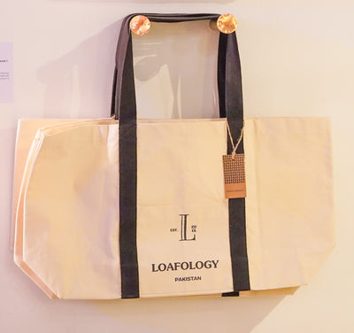 A large, beige Loafology Canvas Tote Bag (Premium) with black handles hangs on a wall. The bag features "Loafology Bakery & Café" printed on it, accompanied by an "L" logo and "EST 2014." A paper tag is attached to one handle.