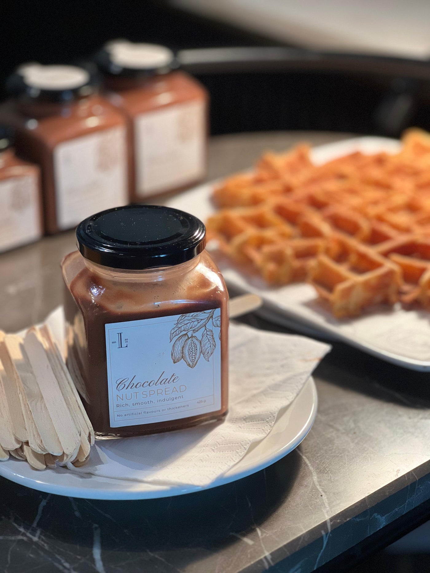 A jar of Choco Nut Spread by Loafology Bakery & Café, free from artificial additives, sits on a plate with wooden sticks in front of plates filled with waffles.