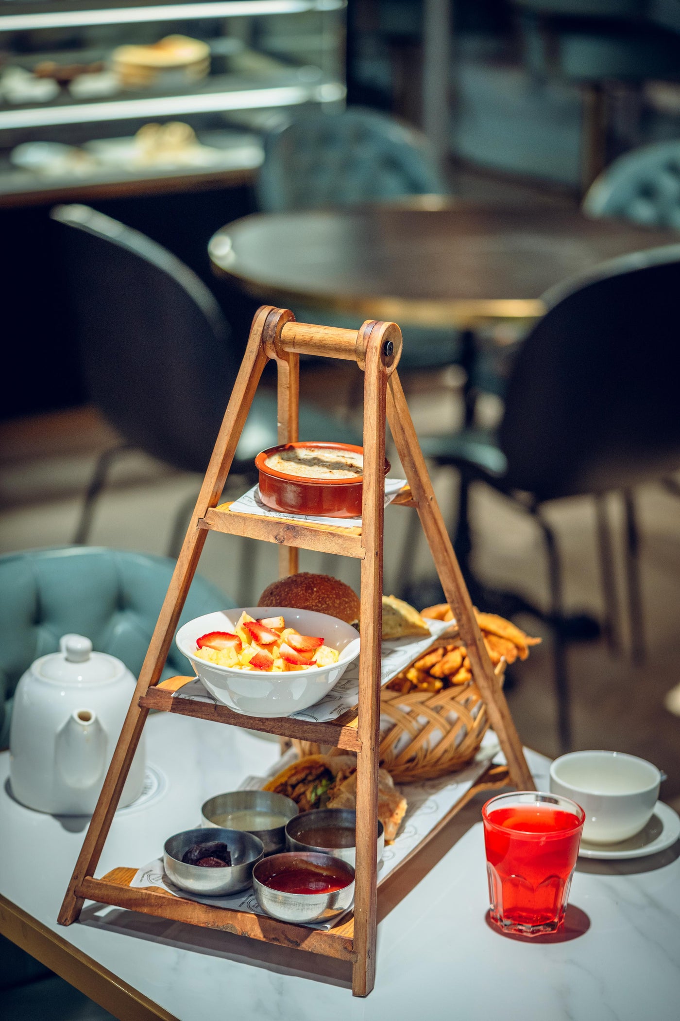 The IFTAR Platter by Loafology Bakery & Café graces the table with a wooden tiered stand of assorted treats, featuring a white teapot, an empty cup, and a red drink in a glass. Among the delights are savory beef sliders that tempt the palate.