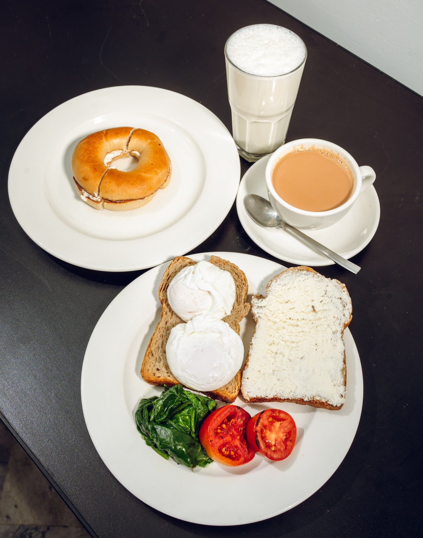 A breakfast spread called "Healthy Sehri" by Loafology Bakery & Café includes a bagel, mug of tea, glass of milk, and toast with poached eggs, fresh spinach lassi, sliced tomato, and cream cheese, all set on a dark table.
