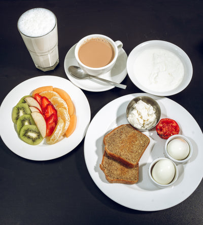 Breakfast on a dark table includes cut fruit, Keto Sehri almond bread from Loafology Bakery & Café, boiled eggs, creamy yogurt, a steaming cup of tea, and a cool glass of milk.