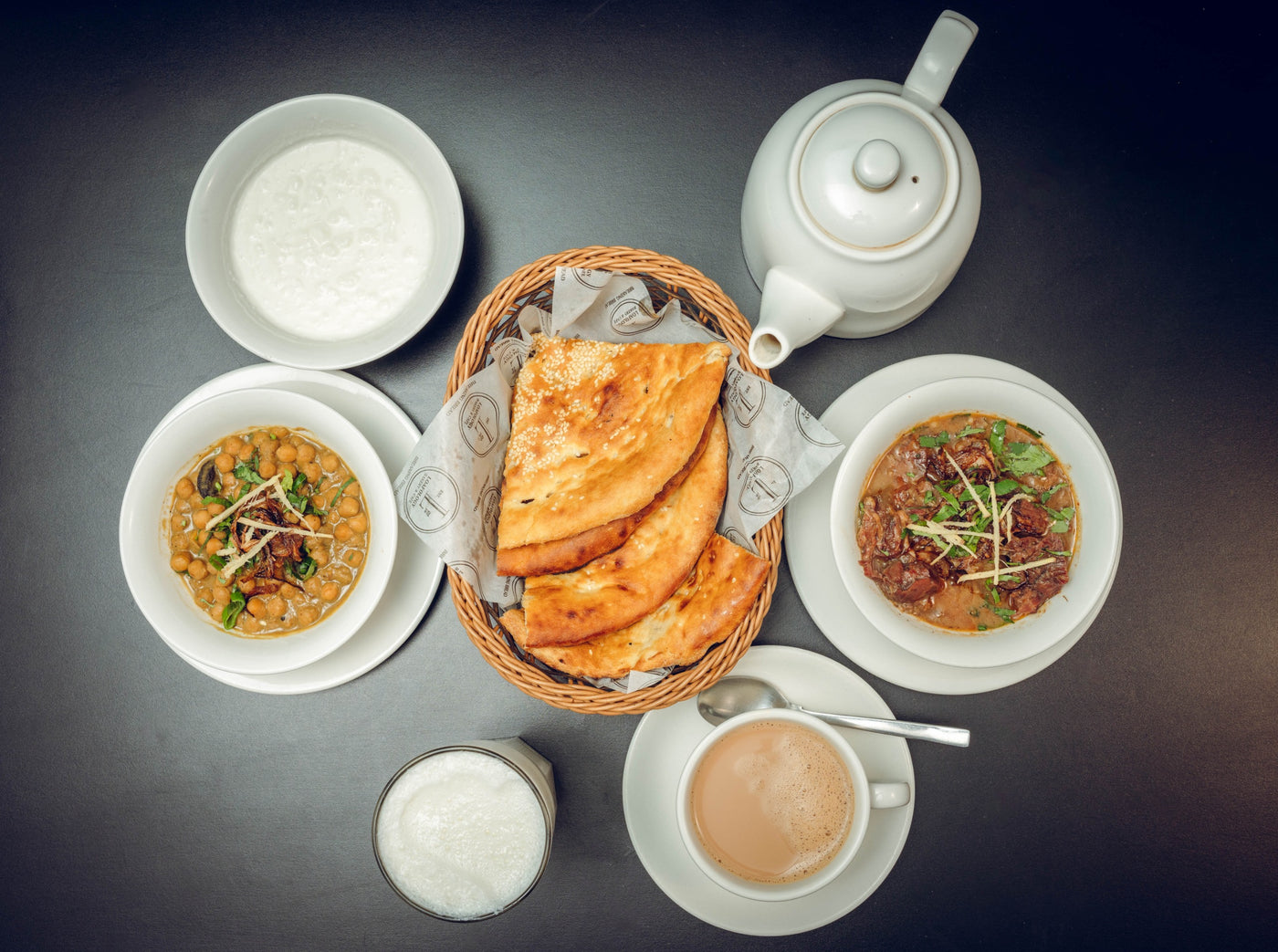 The Desi Sehri 1 set from Loafology Bakery & Café features a table with naan, lentil soup, meat curry, Lahori Channay, yogurt, lassi, and tea in a teapot. These are elegantly arranged on white plates against a sleek black surface.