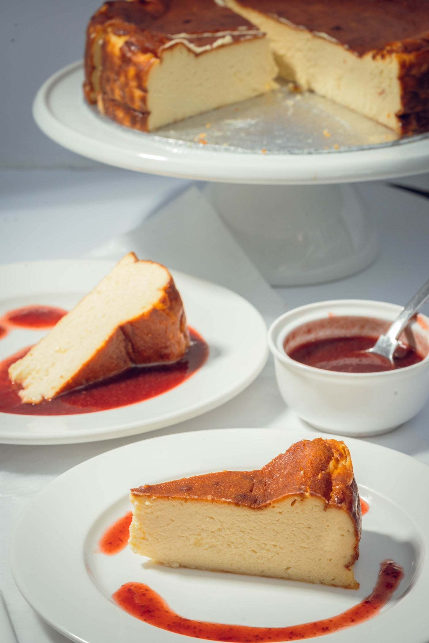 Two slices of Loafology Bakery & Café's San Sebastián Cheese Cake are topped with red sauce on white plates. In the background, a whole cheesecake rests elegantly on a stand, while a nearby bowl of sauce waits with a spoon for the perfect finishing touch.