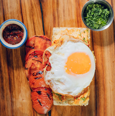 The "Hummus Among Us" from Loafology Bakery & Café features a sunny-side-up egg, grilled tomatoes, and herbs on Focaccia, presented on a wooden surface with red sauce and chopped herbs.