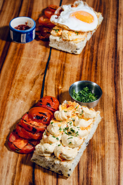 On a wooden surface are two focaccia slices from Loafology Bakery & Café: one topped with grilled tomatoes and herbs, the other with a fried egg. A side of herbs and "Hummus Among Us" is included.