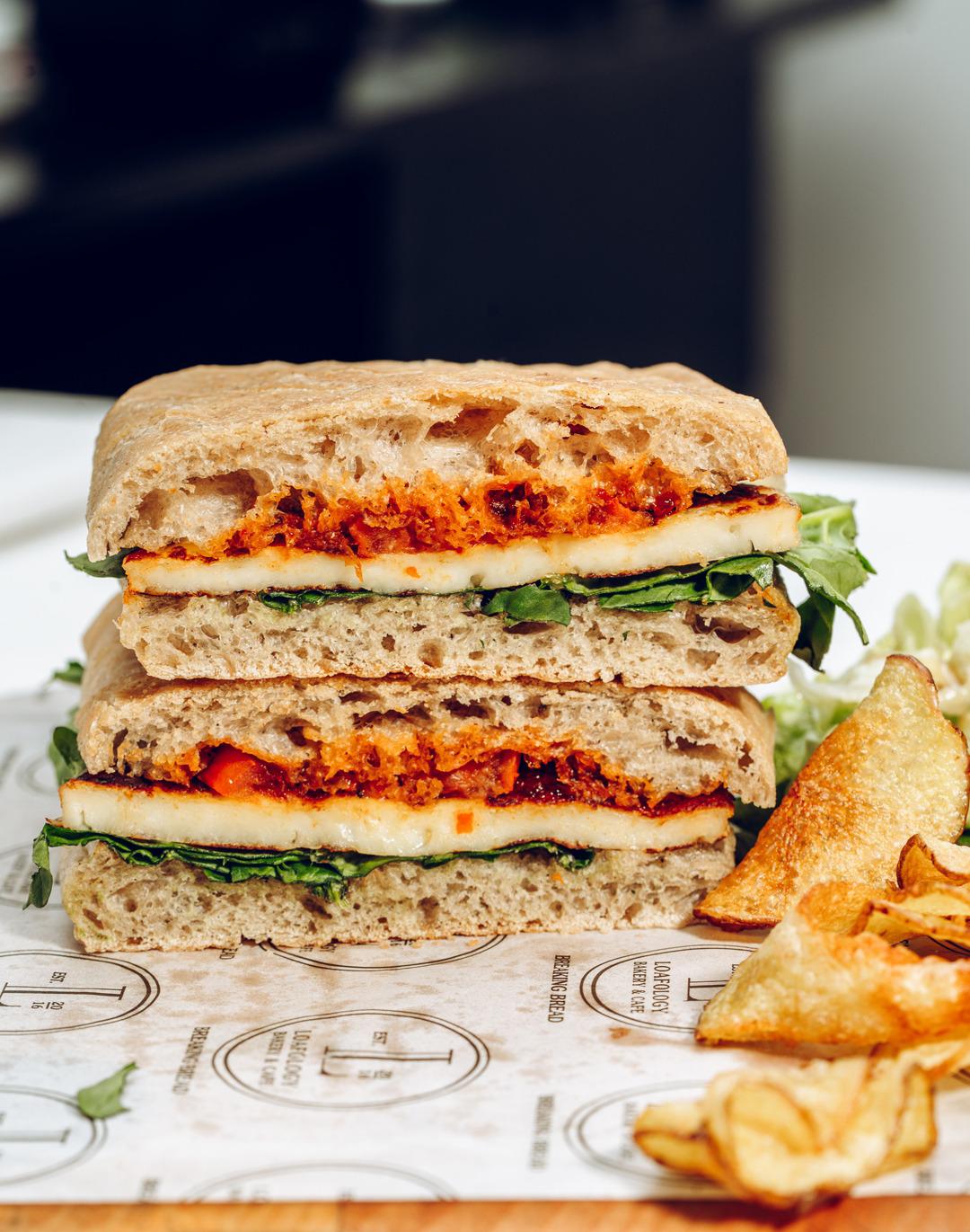 The Halloumi Sizzle from Loafology Bakery & Café features grilled veggies and Halloumi cheese on focaccia, cut and stacked, served on printed paper with potato chips.