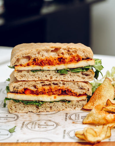 The Halloumi Sizzle from Loafology Bakery & Café features grilled veggies and Halloumi cheese on focaccia, cut and stacked, served on printed paper with potato chips.