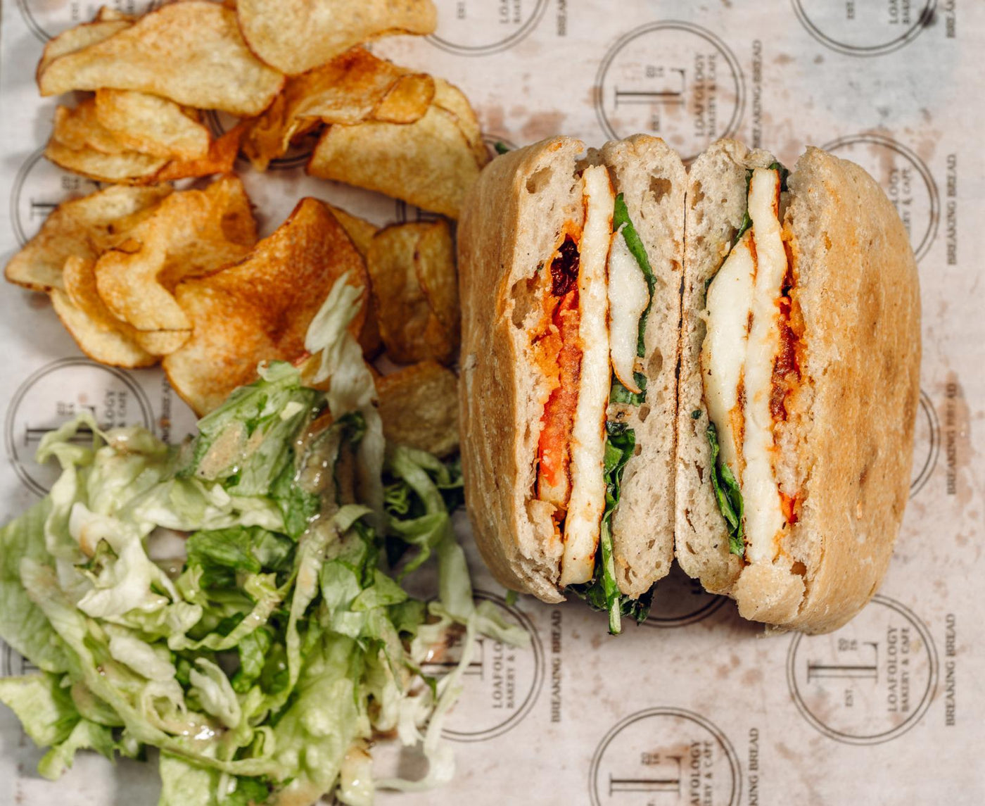 The Halloumi Sizzle from Loafology Bakery & Café is a panini filled with halloumi cheese, spinach, and tomato on ciabatta, served with potato chips and shredded lettuce.