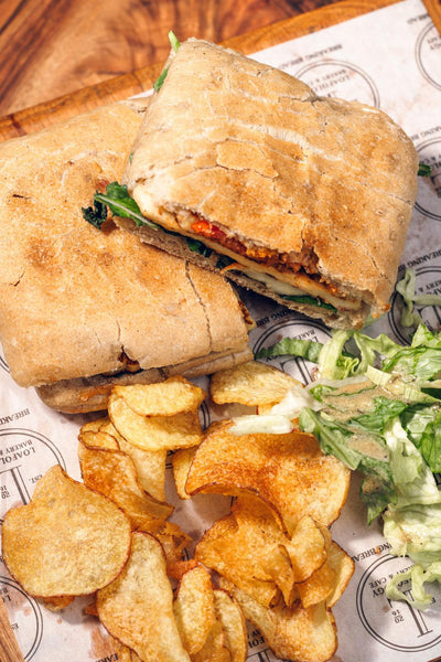 The Halloumi Sizzle from Loafology Bakery & Café is a toasted ciabatta sandwich featuring lettuce, meat, and a touch of chilli jam, accompanied by potato chips and a side of shredded lettuce on paper.