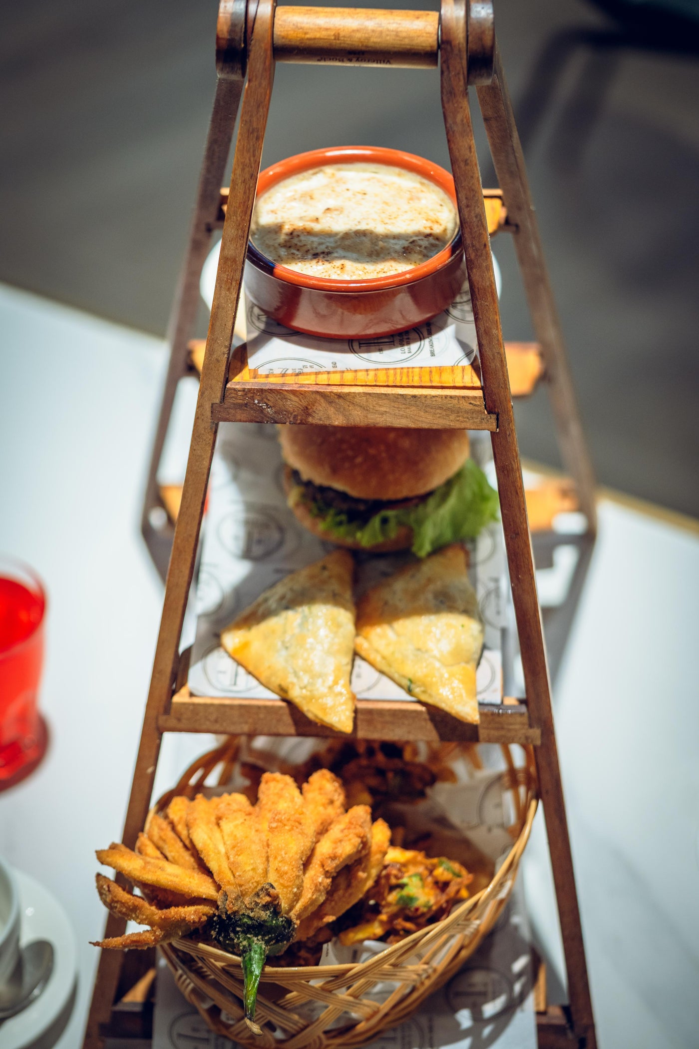 The IFTAR Platter from Loafology Bakery & Café features a charming three-tiered stand with a top small dish, a middle tier Beef Slider with all the fixings, and crispy Vegetable Pakoras in a basket at the bottom.