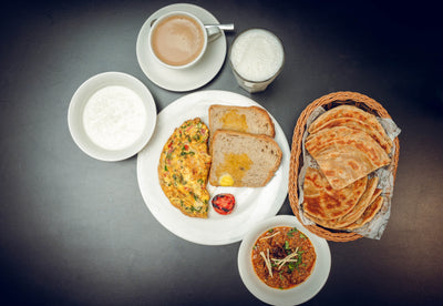 The Desi Sehri 2 breakfast spread by Loafology Bakery & Café features a Desi Omelette, toast, grilled tomato, flatbreads basket, curry bowl, porridge, and refreshing drinks like tea and frothy Lassi on a dark table.