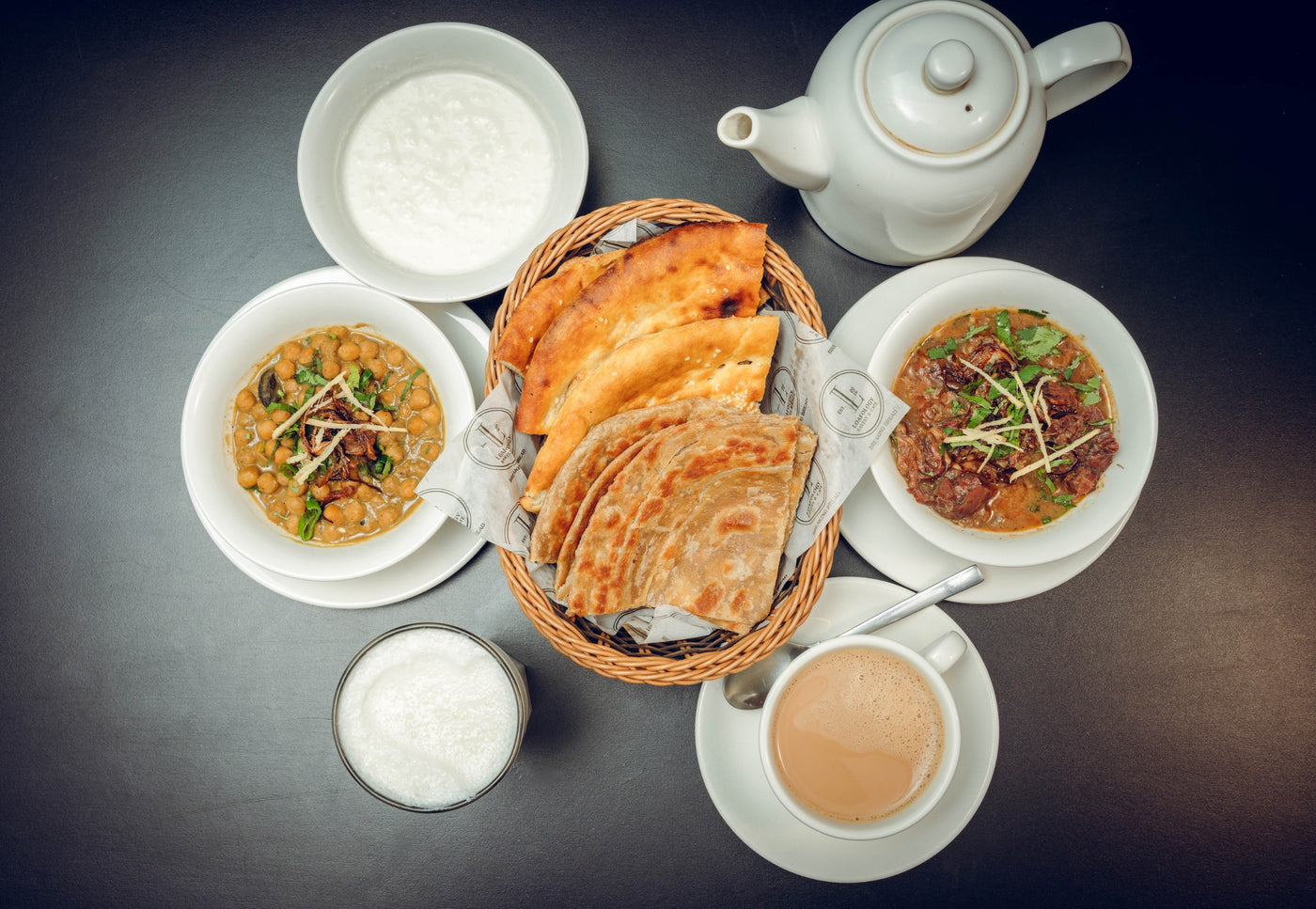 Loafology Bakery & Café's Desi Sehri 1 showcases steaming Parathay, two curry bowls, a hearty porridge bowl, a cup of tea with its teapot, and a refreshing glass of milk elegantly arranged on a dark surface.