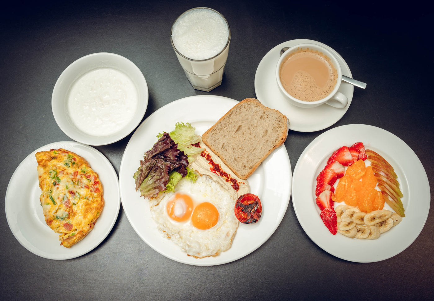 The Loafology Sehri from Loafology Bakery & Café includes a two-egg sunny-side-up, toast, omelette, fruit, yogurt, a glass of milk, and coffee on white plates with a dark table setting, enhanced by a flavorful hummus bowl.