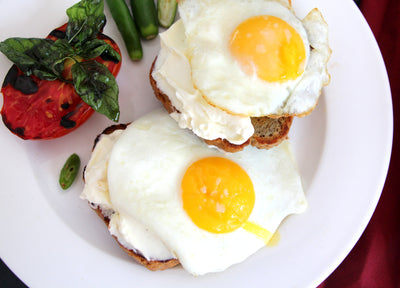 A white plate features Loafology Bakery & Café's Keto Breakfast, showcasing two sunny-side-up eggs on keto almond bread, accompanied by a roasted tomato, green pepper, and basil leaves for an ideal low-carb morning meal.