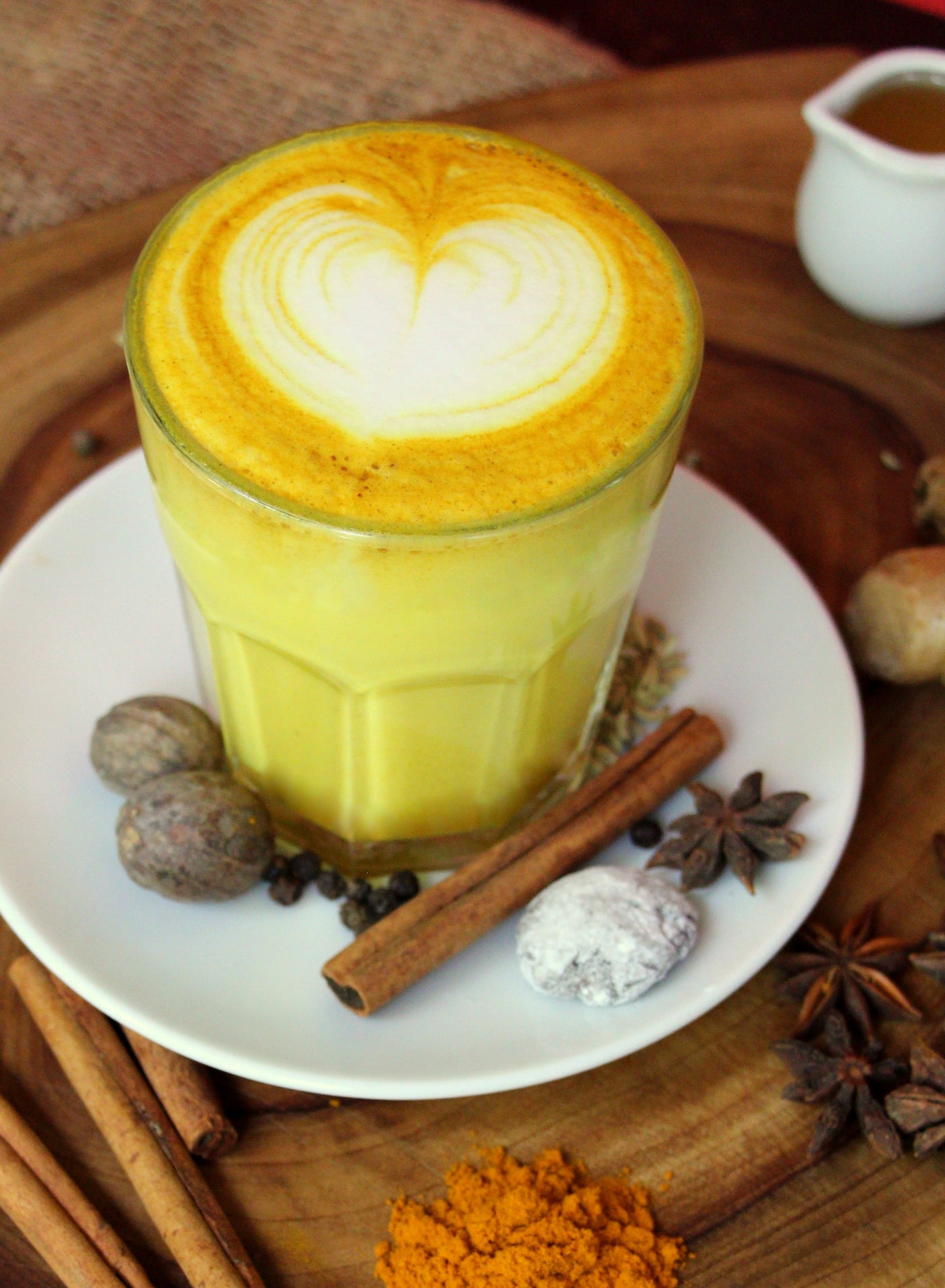 A glass of Loafology Bakery & Café's Turmeric Latte on a white plate, surrounded by spices including cinnamon sticks, nutmeg, star anise, and turmeric powder, creates a vibrant health drink on a wooden surface.