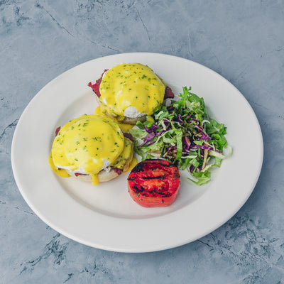 Introducing the "Beef Bacon Benedict" by Loafology Bakery & Café: A delectable plate featuring eggs Benedict drizzled in rich hollandaise sauce, served with crispy beef bacon, a perfectly grilled tomato, and a petite side salad composed of fresh greens and shredded vegetables, all beautifully presented on a gray background.