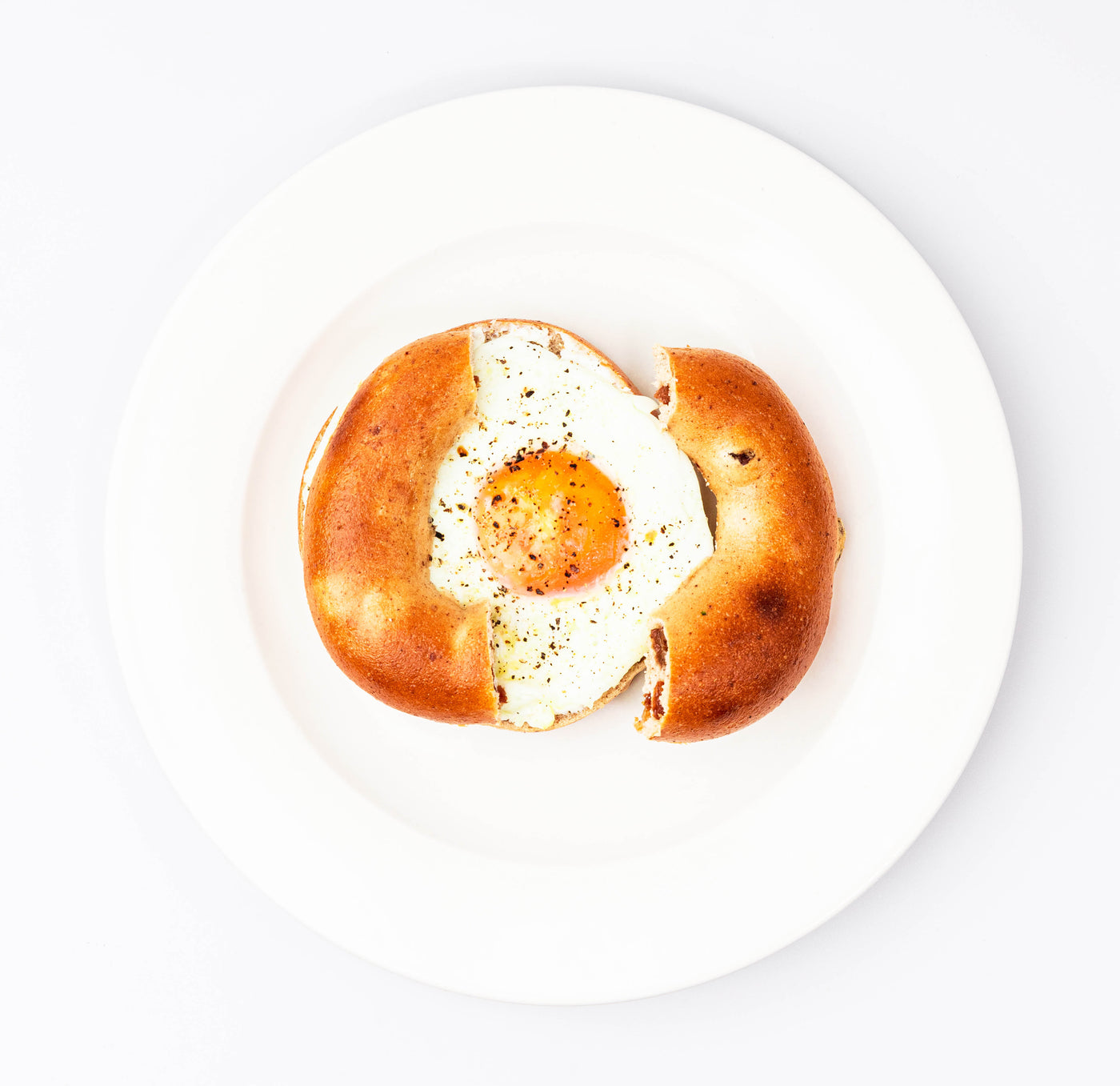 A sunny-side-up egg cooked in the center of a halved Breakfast Fix bagel, spread with cream cheese, on a white plate from Loafology Bakery & Café.