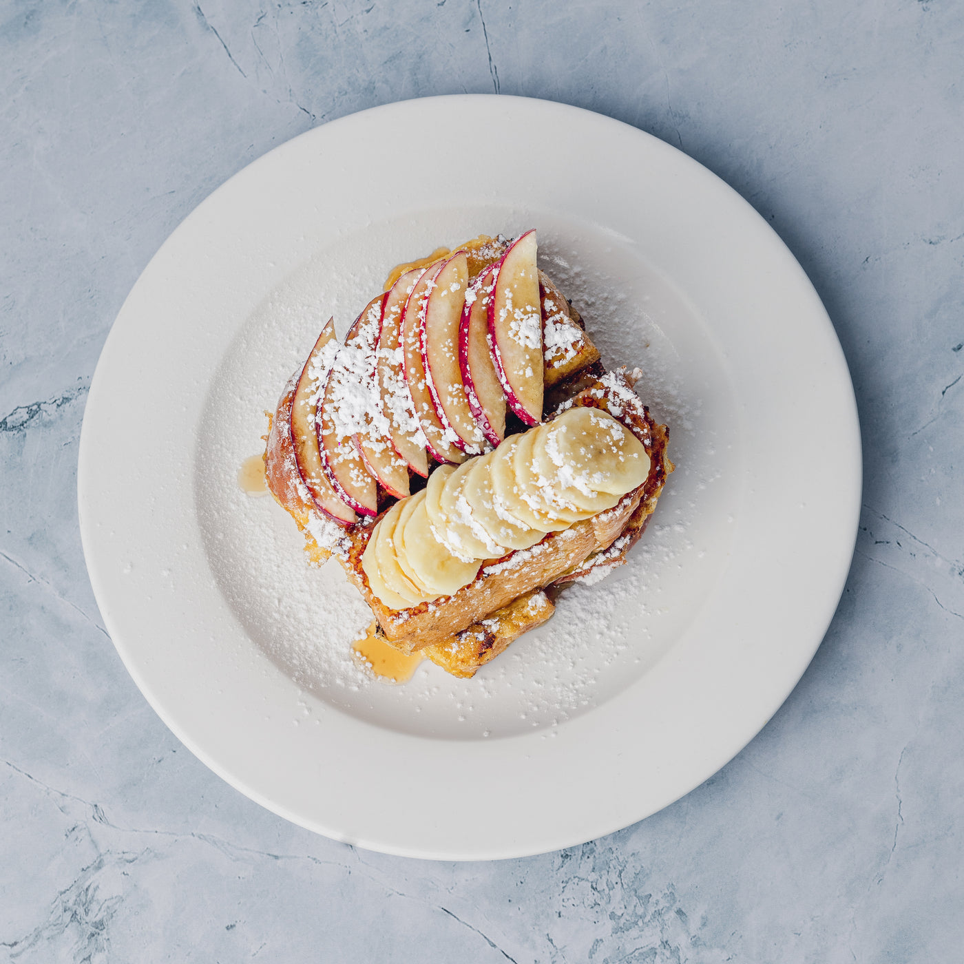 A white plate featuring Loafology Bakery & Café's Brioche French Toast, adorned with powdered sugar, banana slices, and thinly sliced seasonal fruit, set against a light gray marble surface.