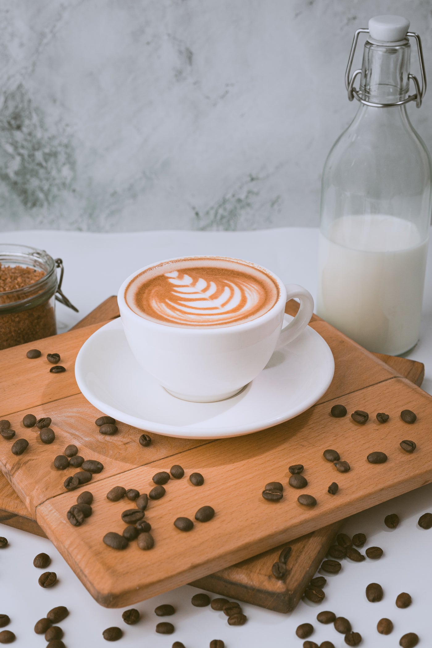 A cup of Loafology Bakery & Café's Decaffeinated Coffee with latte art on a white saucer, placed on a wooden board with scattered coffee beans, a glass jar of sugar, and a glass bottle of milk in the background. Enjoy this caffeine-free delight that's as beautiful as it is soothing.