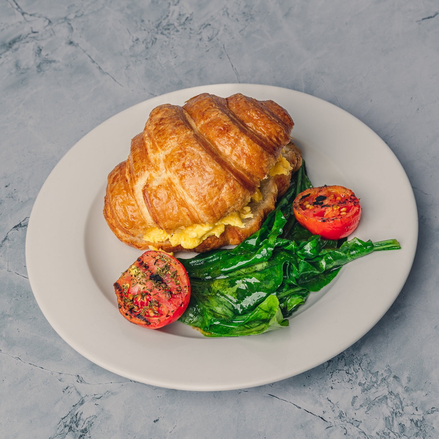 A Toasted Stuffed Croissant from Loafology Bakery & Café, filled with organic scrambled eggs, served with sautéed spinach and grilled tomato halves on a white plate.