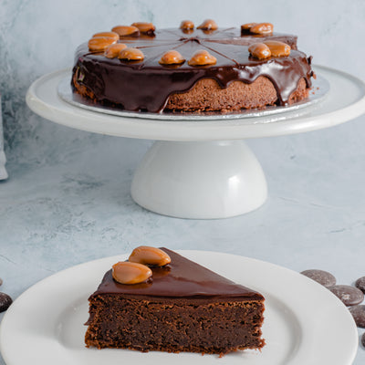 A whole Dark Chocolate & Almond Cake from Loafology Bakery & Café with caramel toppings on a white cake stand and a slice of the cake on a white plate in the foreground.