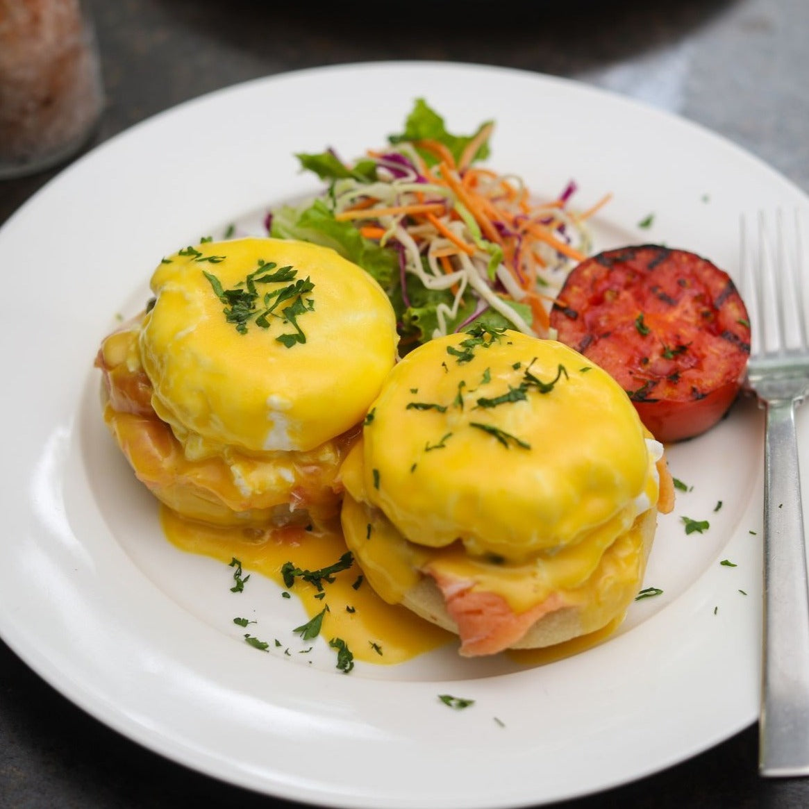 A plate of Loafology Bakery & Café's Eggs Royale features organic poached eggs and hollandaise sauce, garnished with herbs, served alongside a small salad and a grilled tomato.