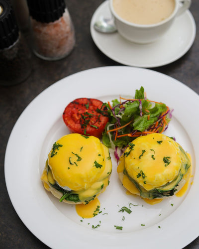 A plate of Eggs Florentine featuring organic poached eggs with creamy hollandaise sauce, garnished with herbs, and served alongside a grilled tomato, wilted spinach, and a side salad from Loafology Bakery & Café. A cup of coffee and salt and pepper shakers are in the background.