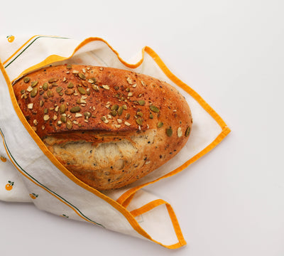 A loaf of Loafology Bakery & Cafe's Eight Grain Bread, partially wrapped in a white cloth accented with an orange border and small orange designs, set against a white background.