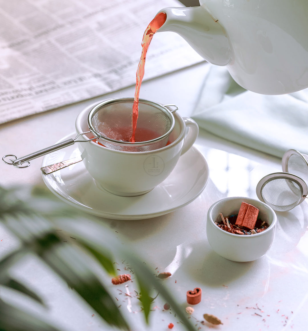 Cardamom Nights Tea by Loafology Bakery & Café is being poured into a teacup through a strainer, releasing a soothing aroma that eases muscles. In the background, a small bowl of tea leaves and a newspaper set the perfect scene for relaxation.