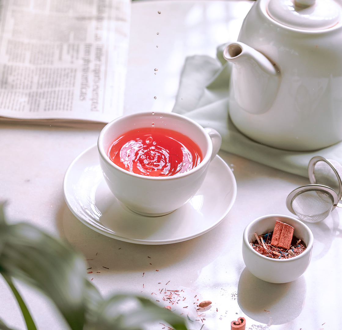 A cup of Loafology Bakery & Café's Cardamom Nights Tea rests on a white saucer, accompanied by a tea kettle, a small bowl of tea leaves, a tea strainer, and part of a newspaper in view. This soothing blend is perfect for unwinding after a long day and may help soothe muscles with its anti-inflammatory properties.