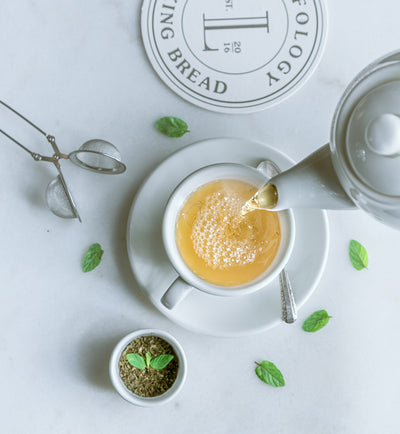 A Loafology Bakery & Café Peppermint Tea teapot pours tea into a cup on a saucer, surrounded by refreshing peppermint leaves. A small dish with loose tea and a caffeine-free infuser is nearby on a light surface.