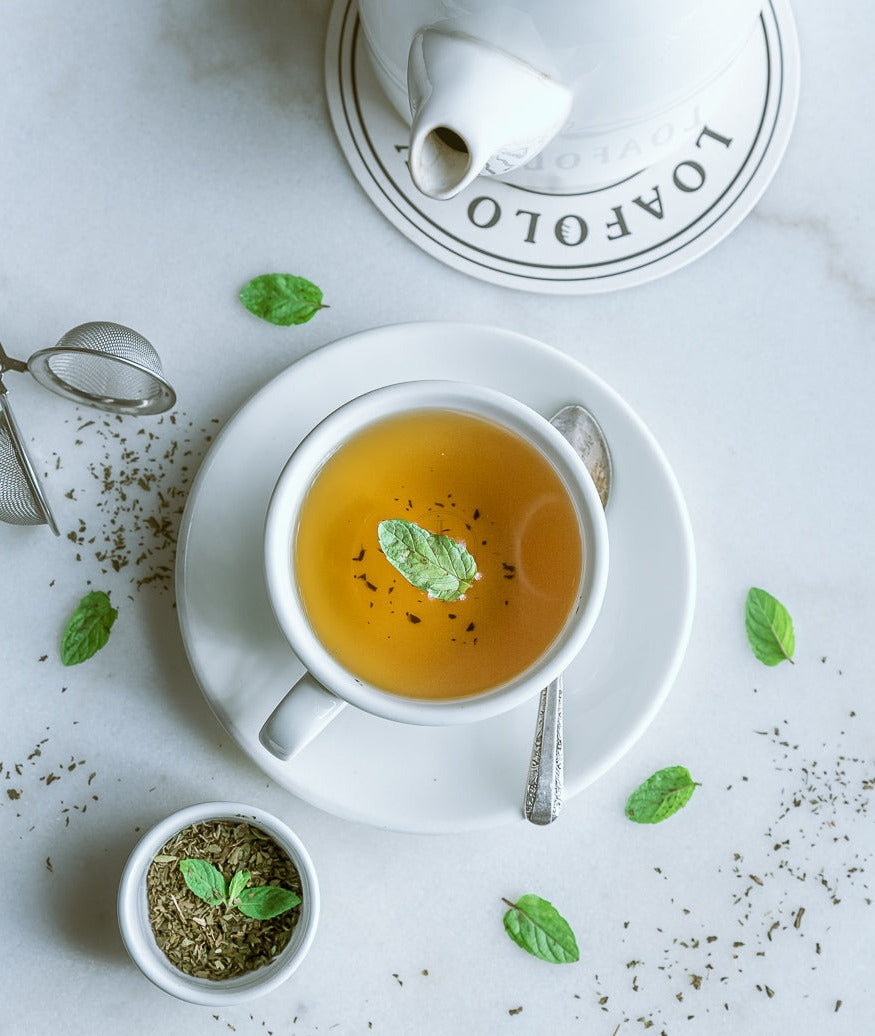 A cup of Loafology Bakery & Café's Peppermint Tea, topped with peppermint leaves, rests on a saucer and is surrounded by loose tea leaves and mint sprigs. A caffeine-free tea bag infuser and a teapot are nearby on the marble surface.