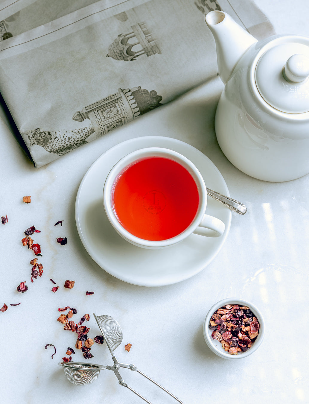 A cup of Loafology Bakery & Café's Strawberry & Kiwi Tea rests on a saucer on the table, accompanied by a white teapot, loose tea leaves, and a strainer. In the background, a newspaper featuring architectural images peeks through, creating an ideal setting for your evening tea ritual.